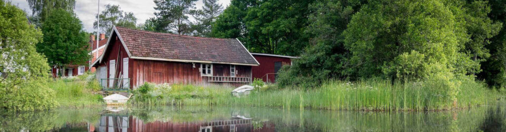 Traditionellt rött trähus vid en lugn sjö med reflektioner i vattnet och omgivet av grönska.